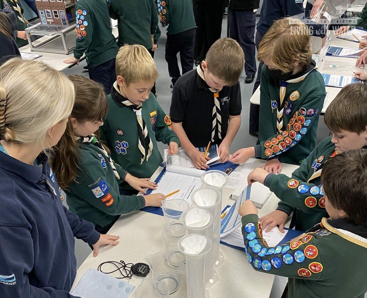An image of six scouts huddled around a table taking part in an activity, with a scout leader overseeing.